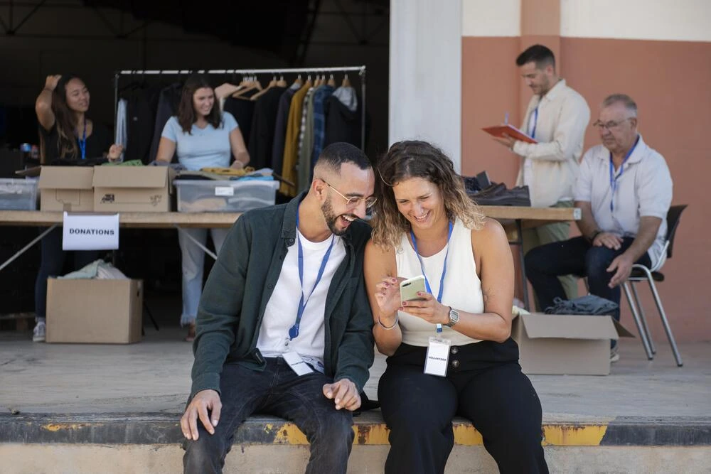 A group of people are gathered at a table with boxes labeled "DONATIONS." Two people in the foreground are looking at a phone and laughing. Others are working at the table, sorting clothes. The setting appears to be an outdoor location.