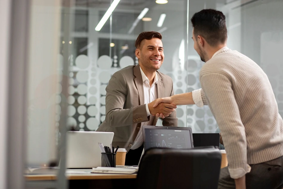 Two men are shaking hands in an office setting. One man is wearing a suit jacket and smiling, while the other is wearing a sweater and facing away from the camera. Laptops and office supplies are visible on the desk.