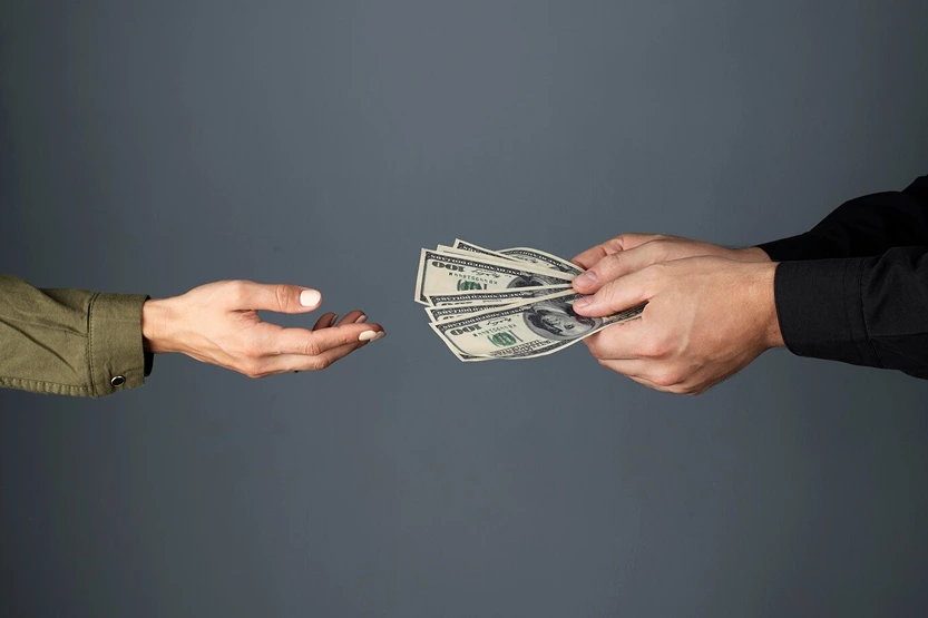 Two hands are shown exchanging several US $100 bills against a solid gray background. One hand is open and reaching out, while the other is holding the money.