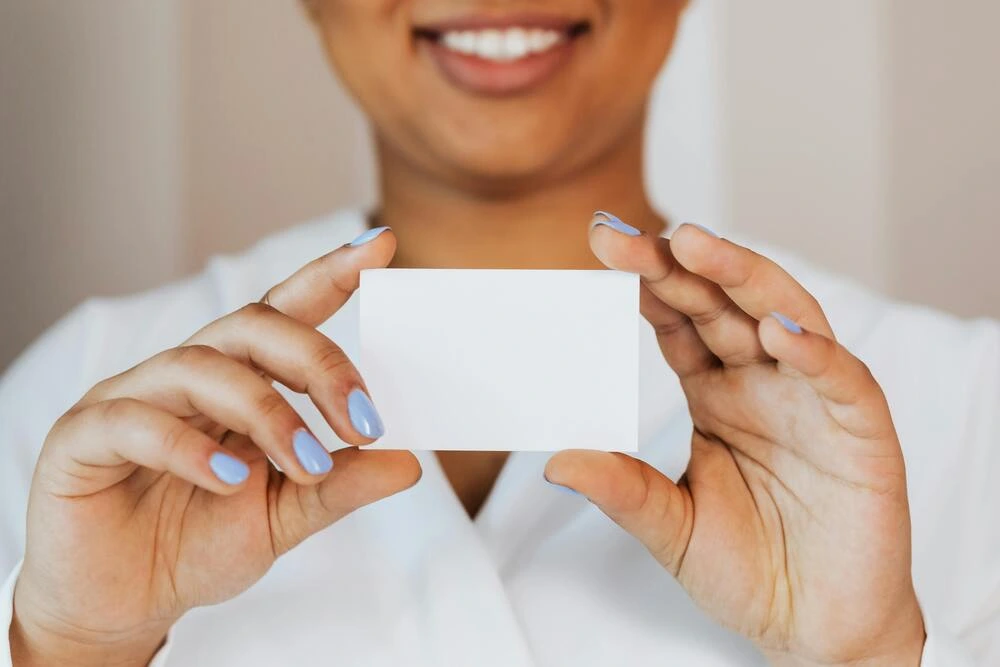 A person with a bright smile holds a blank white card in their hands. The person's face is partially visible, and they have light blue nail polish.