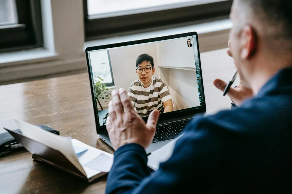 A person with a shaved head is seen from behind, gesturing with their hands while participating in a video call on a laptop. The laptop screen shows another person with dark hair and glasses in a striped shirt.
